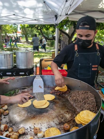 A Tacos Costa cook working a large round plancha of tortillas, grilled onions and meat under a tent at an outdoor event.