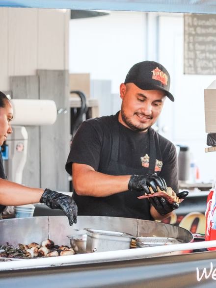 A smiling Tacos Costa cook in a black cap and gloves preparing food inside the truck.