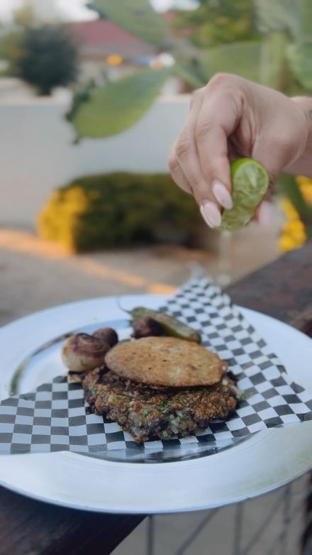 A hand squeezing fresh lime over a plate on checkered paper, with grilled onion and jalapeño on the side.
