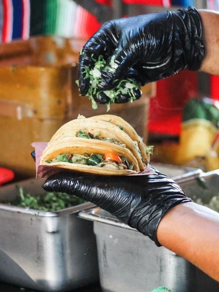 Gloved hands sprinkling chopped cilantro over a pair of folded tacos at the truck.