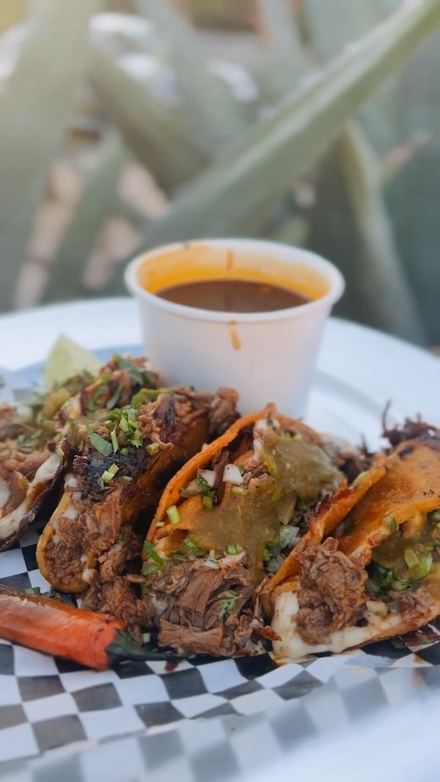 Crispy red-orange tacos topped with green salsa, onion and cilantro, with a cup of consomé on the plate.
